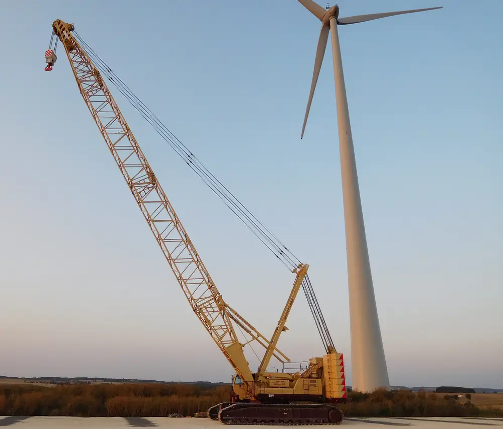 A lattice-boom crawler crane on tracks with its boom raised, standing beside a wind turbine at dusk